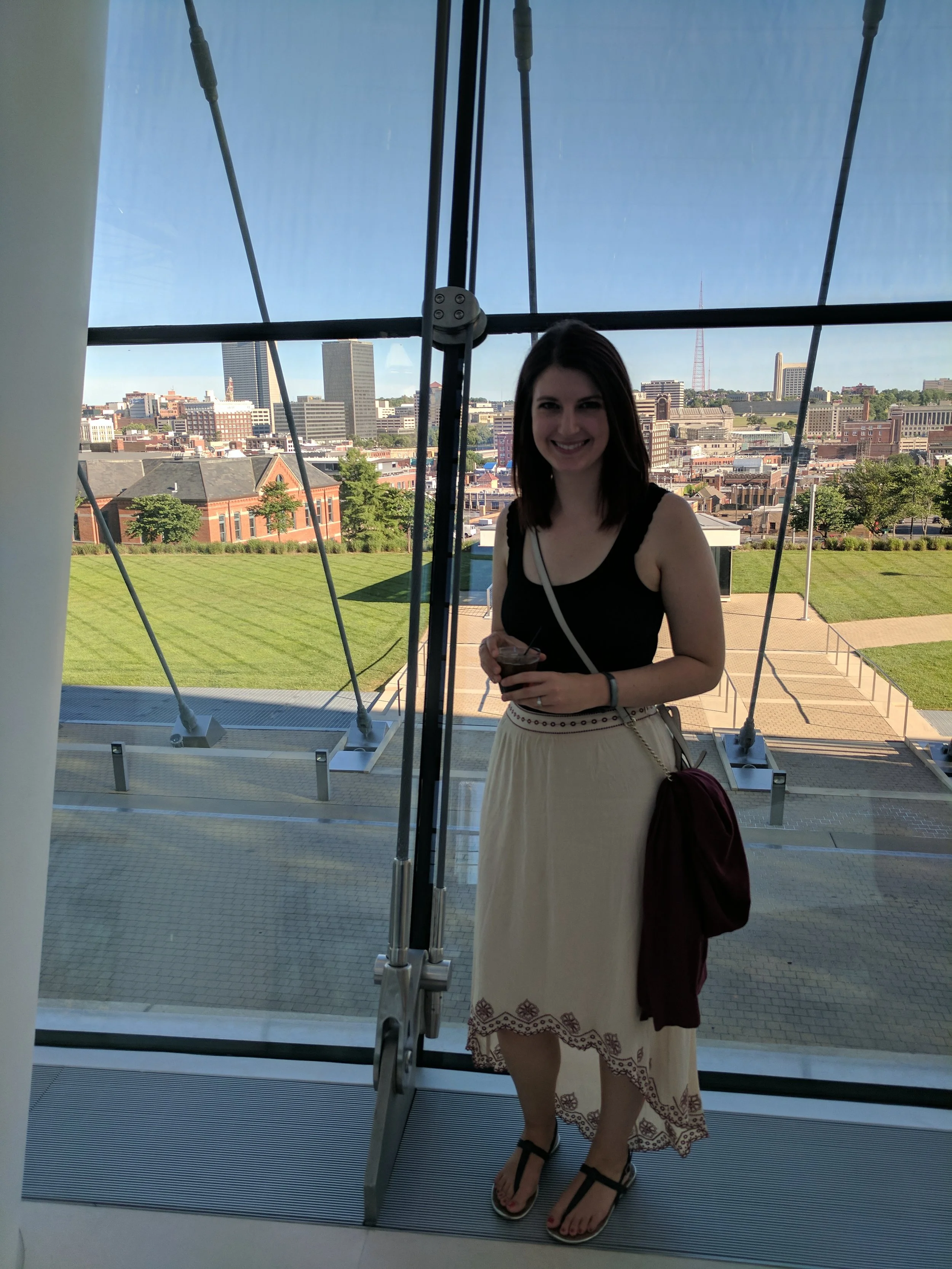 A woman stands inside a building near a glass door, smiling at the camera. She has shoulder-length dark hair and is holding a drink. She wears a black sleeveless top, a cream-colored skirt with embroidered patterns, black sandals, and carries a maroon bag. The background shows a city skyline with various buildings and a clear blue sky.