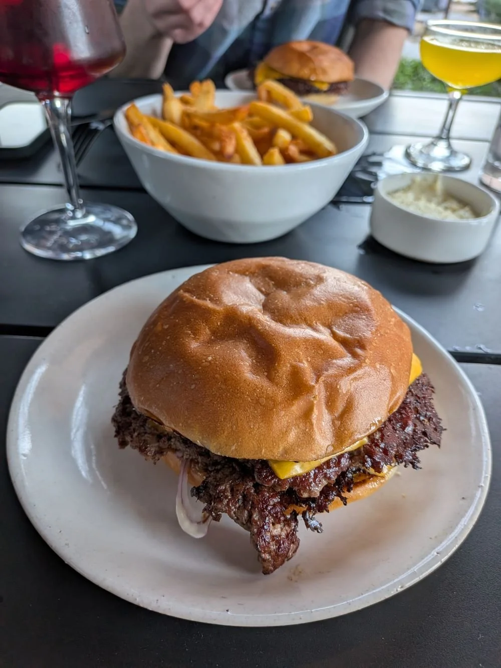 A cheeseburger with a beef patty, cheese, and bun on a white plate. In the background, there is a bowl of French fries, a glass of red wine, and a glass of yellowish cocktail. Also visible are a small bowl of white sauce, another plate with a burger in the distance, and a person's arm at the table.