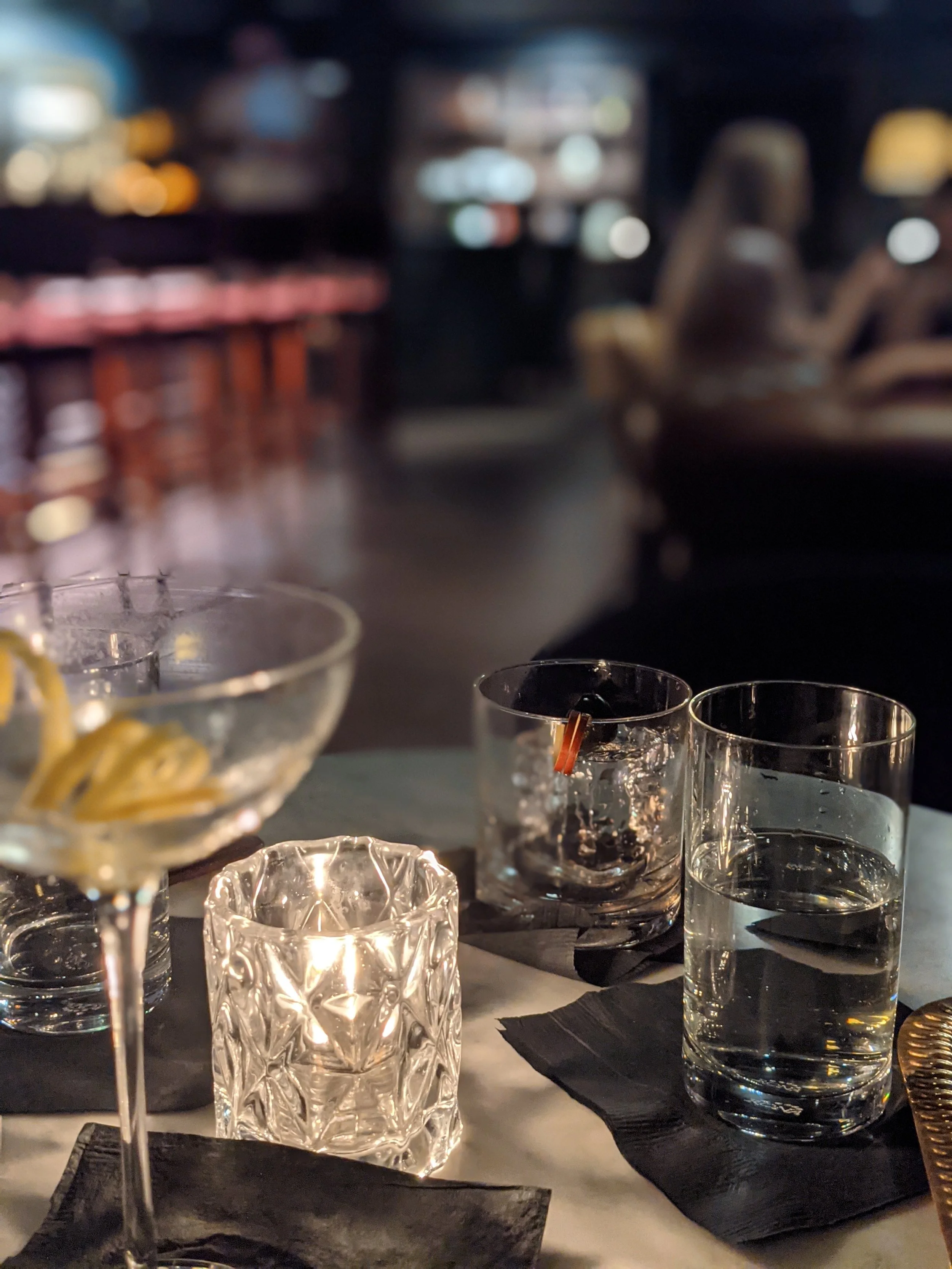 Blurred image of a dimly lit bar or restaurant with glassware on a table, including wine glasses and tumblers, black napkins, and some decorative elements.