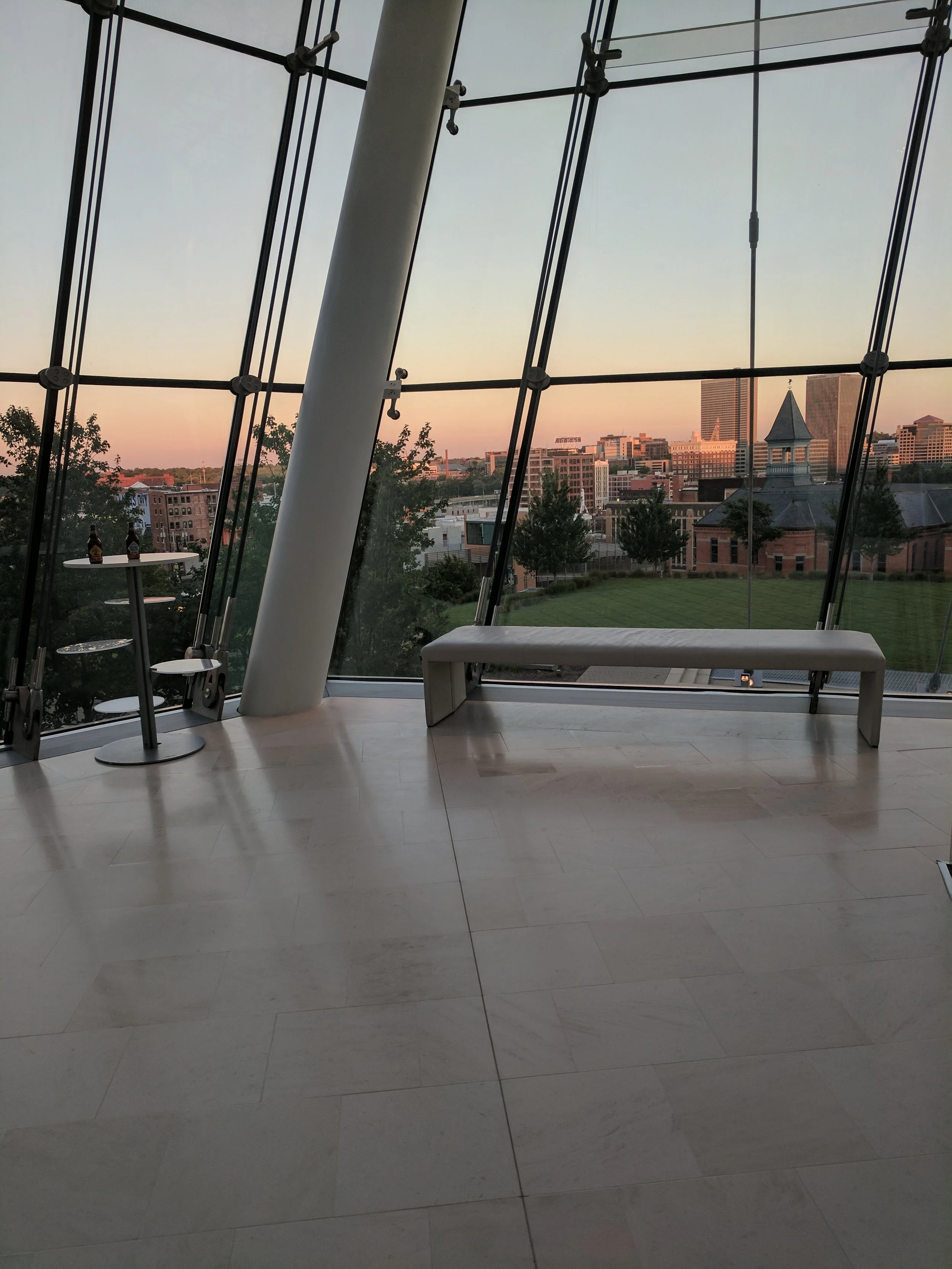 Looking through a large glass window at a cityscape with buildings, trees, and a church with a steeple at sunset. Inside, there are metal chairs and a bench.