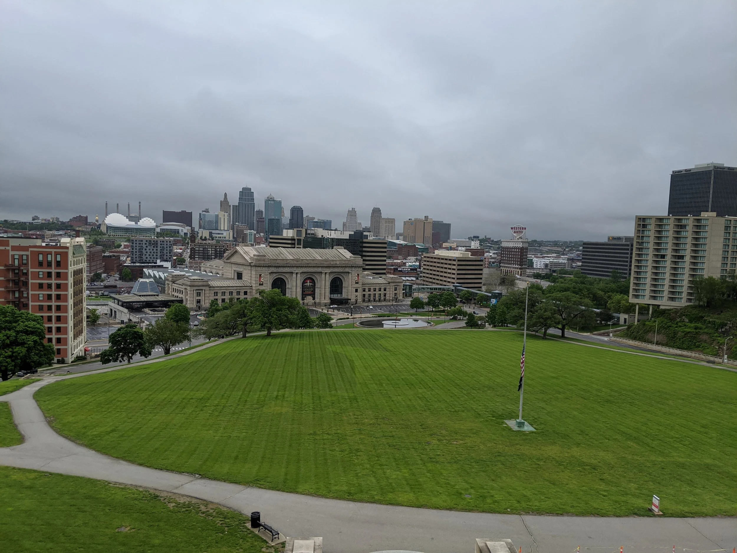 City skyline with tall buildings under cloudy sky, green park in foreground with walking paths and benches.