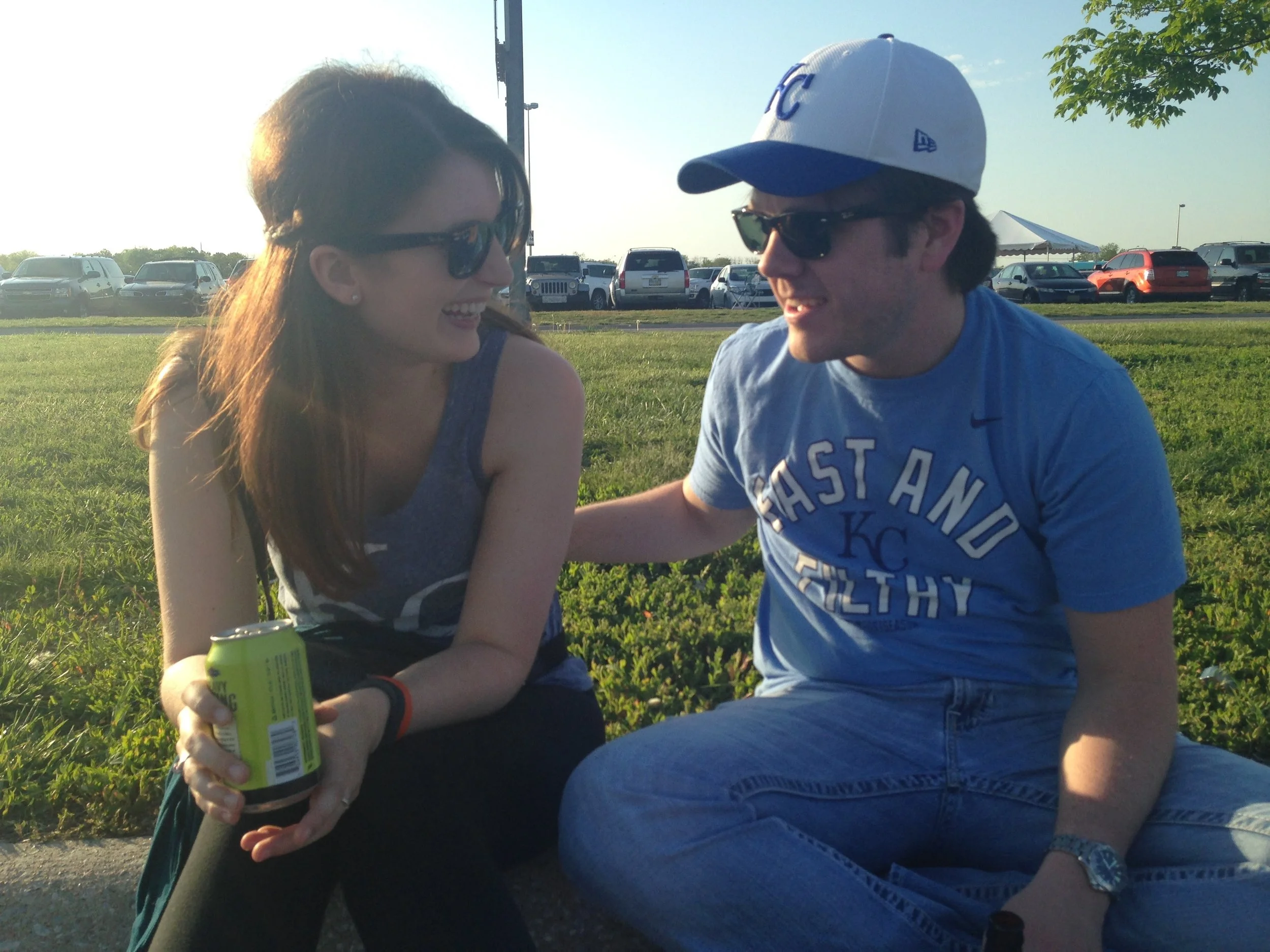 A young woman and a young man sitting outdoors on the grass, smiling and looking at each other, with parked cars and a tree in the background during daytime.
