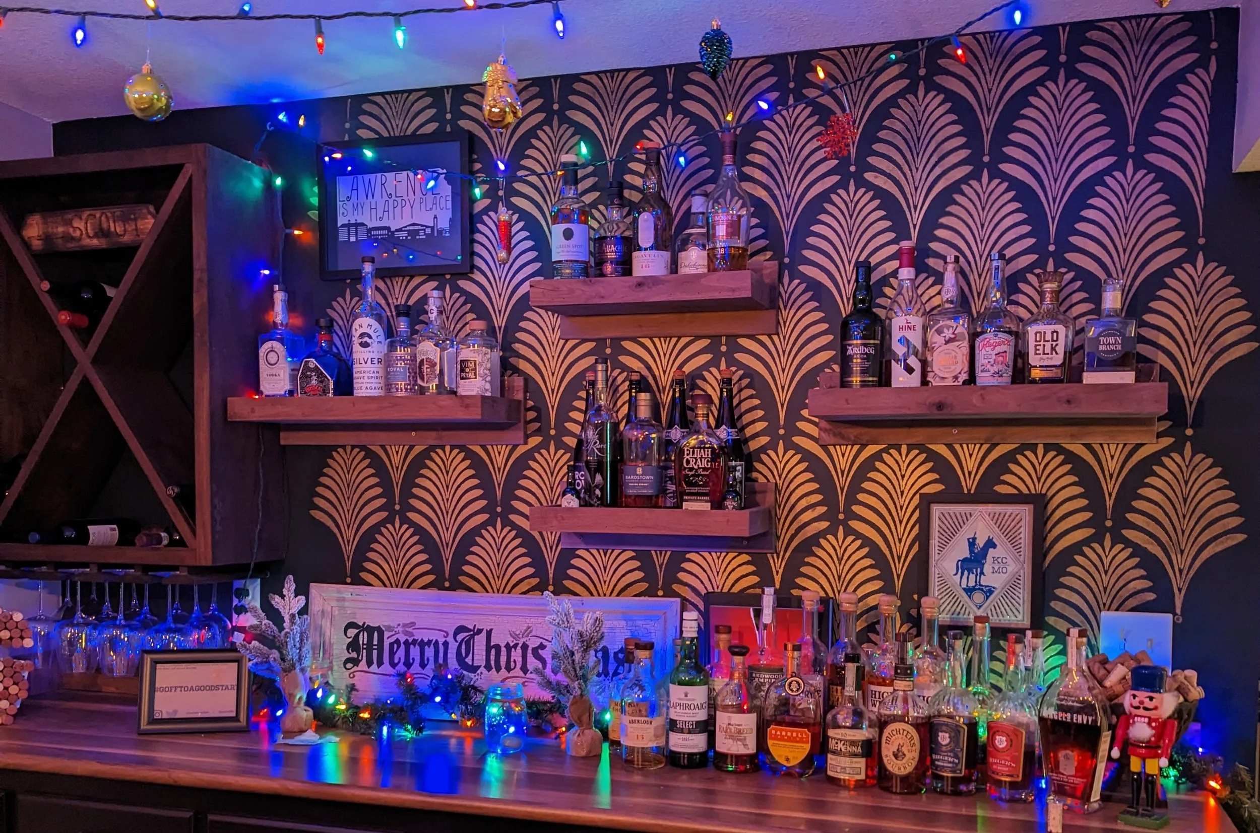 A bar area decorated with colorful Christmas lights, ornaments, and holiday decorations, with shelves of various liquor bottles and a festive 'Merry Christmas' sign.