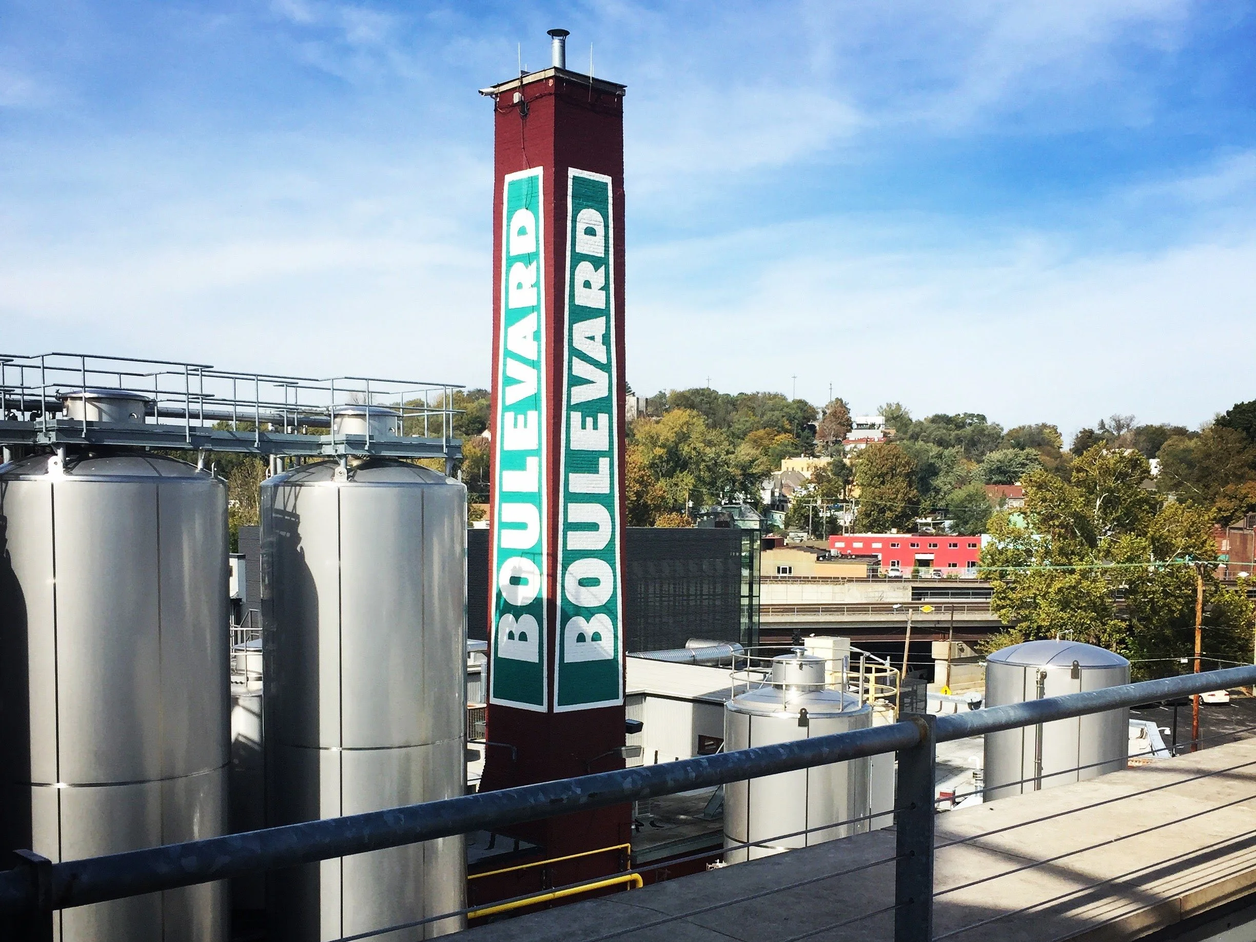 Silver brewery tanks and smokestack with "Boulevard" printed on the side and trees and buildings in the background under a blue sky.