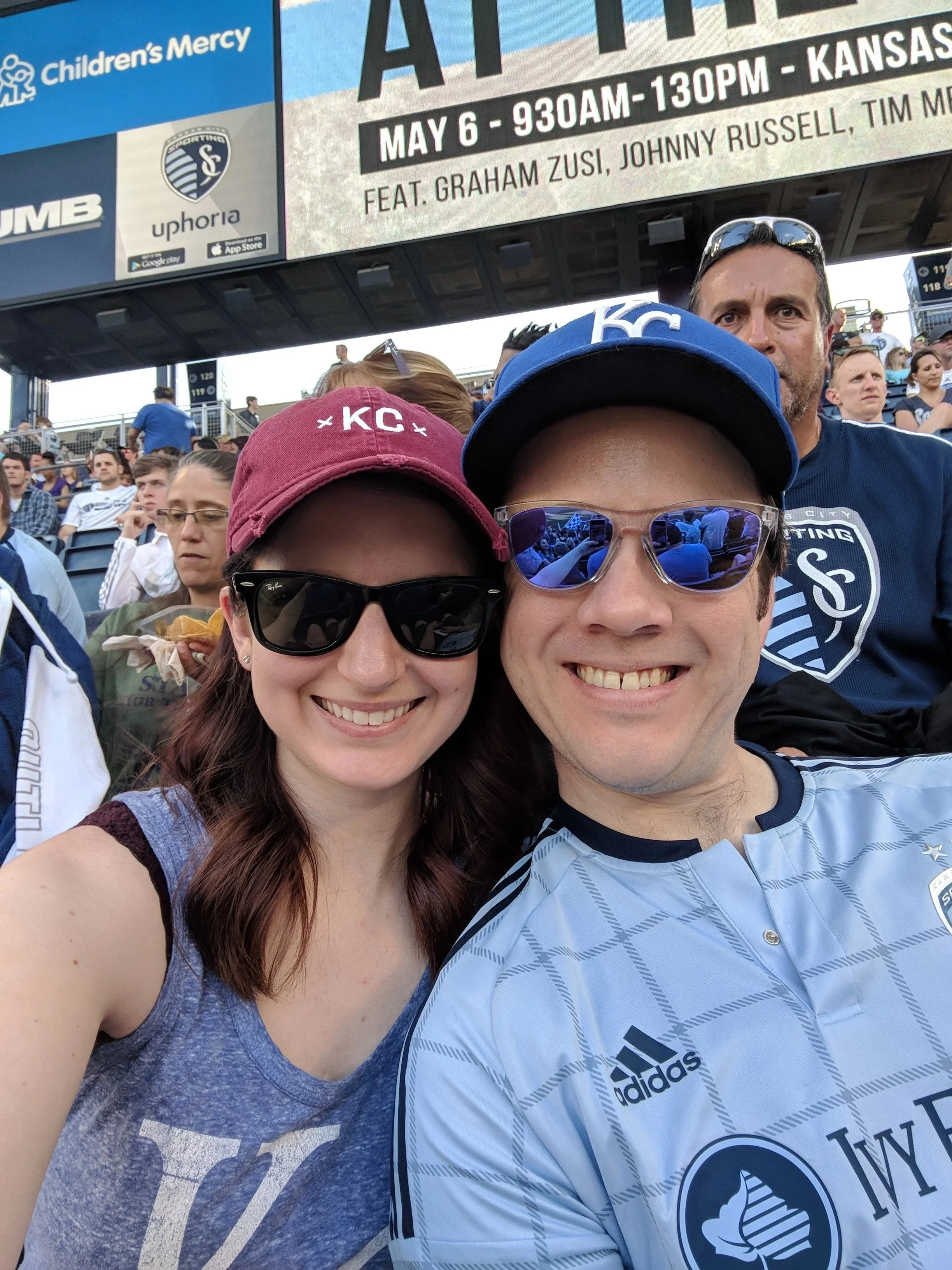 A group of people at a sporting event, with two smiling individuals in the foreground wearing sunglasses and KC caps, and a man in the background wearing a Sporting Kansas City shirt. The background shows a large digital billboard advertising charity event details.