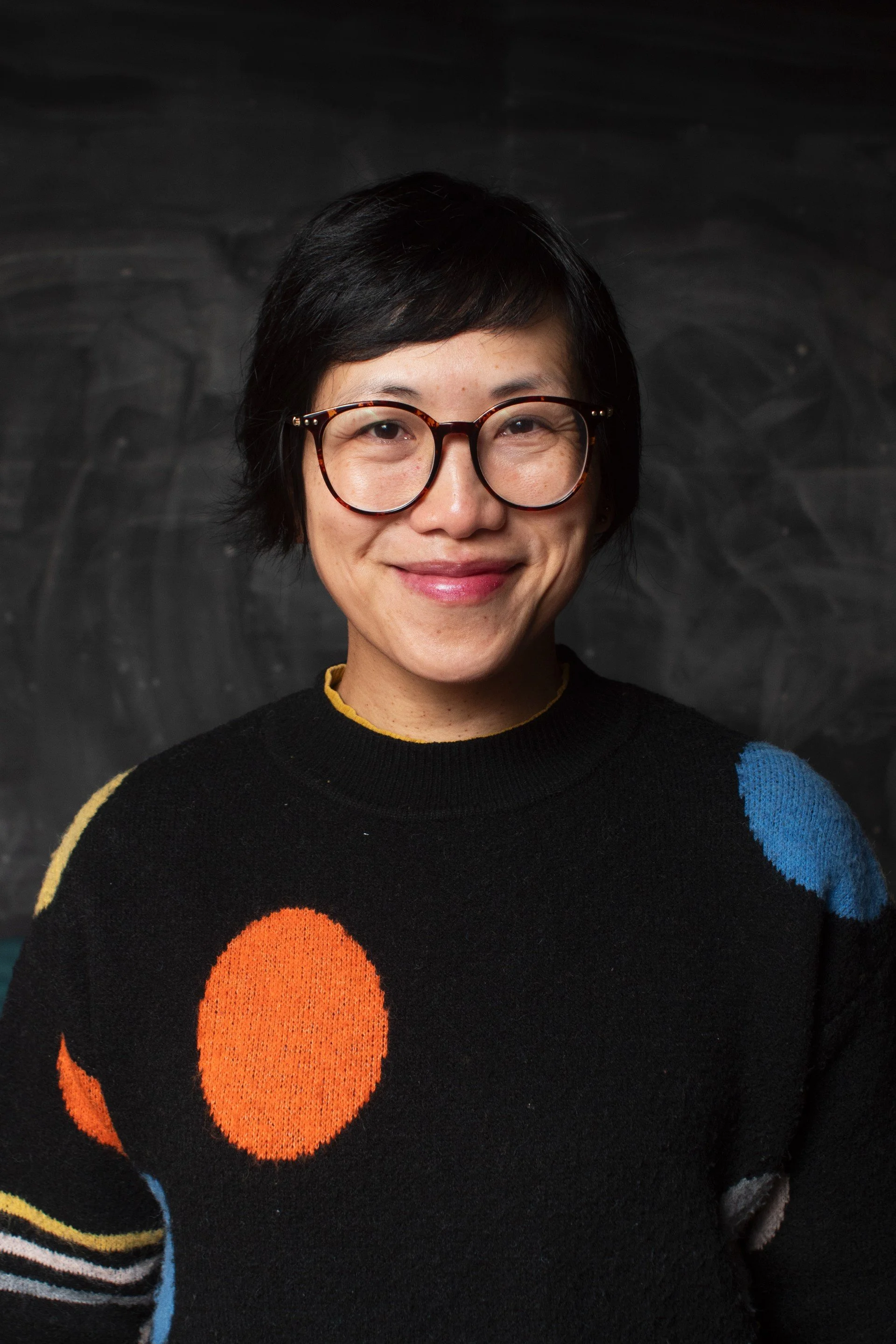 A woman with dark hair in glasses and wearing a polka dot jumper smiles.