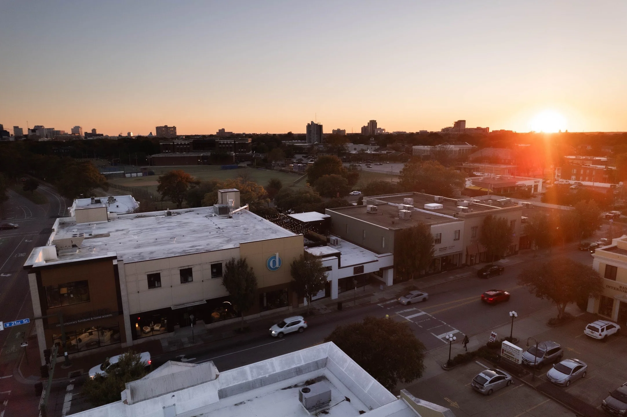 Cityscape at sunset showing buildings, streets, and a park with trees and a sports field.