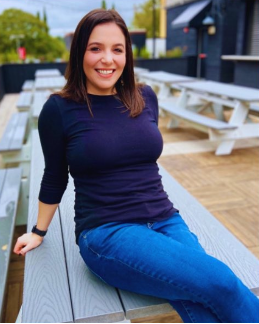 A woman with shoulder-length dark hair smiling and sitting on a wooden picnic table outdoors, with multiple white picnic tables in the background and greenery around.