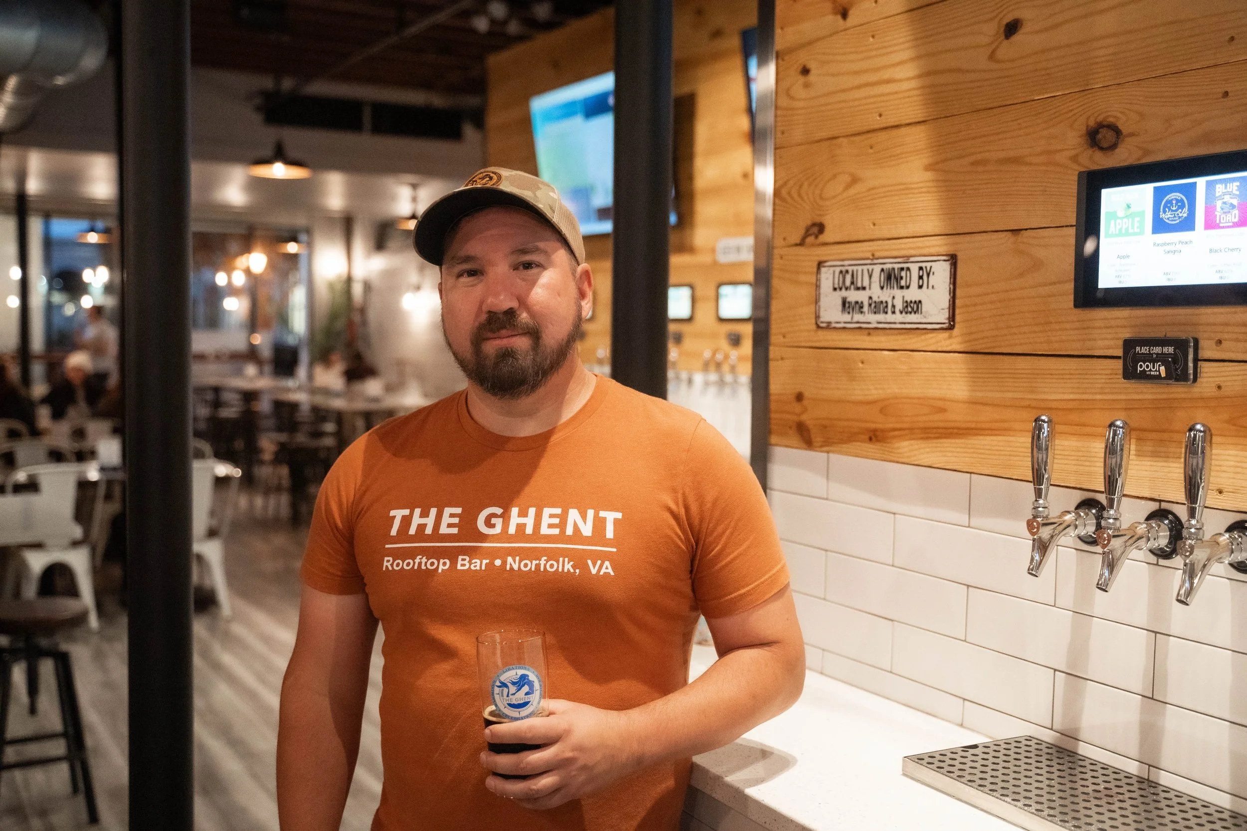 One of the owners with a beard wearing an orange T-shirt that says 'The Ghent Rooftop Bar Norfolk, VA' and a beige cap, holding a glass of dark beer, standing at a bar with wooden paneling and multiple beer taps in a self-pour bar setting.