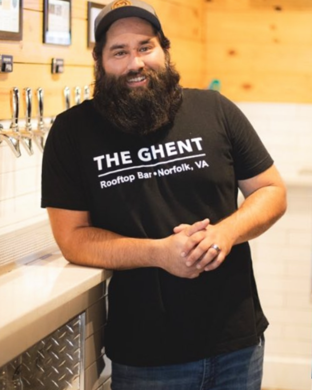 A man with a beard and dark hair wearing a black t-shirt with white text that reads 'THE GHENT Rooftop Bar - Norfolk, VA' stands in a wooden bar or restaurant setting.