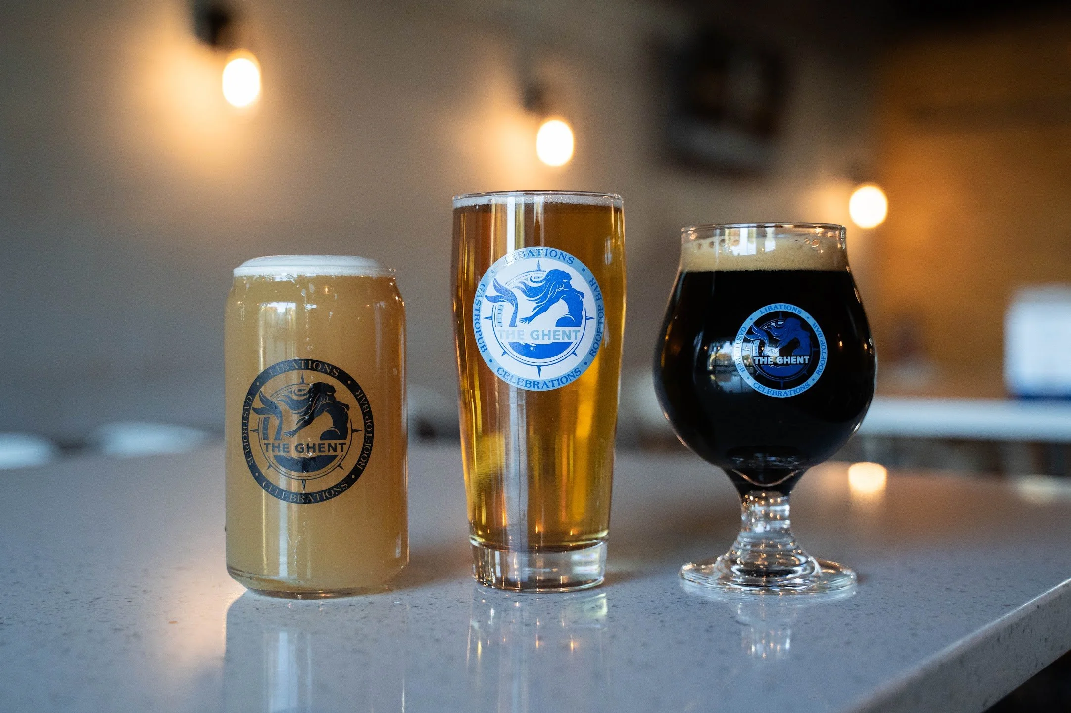 Three glasses of beer on a white countertop, each with a logo sticker, in a bar setting with warm lighting and wooden walls.