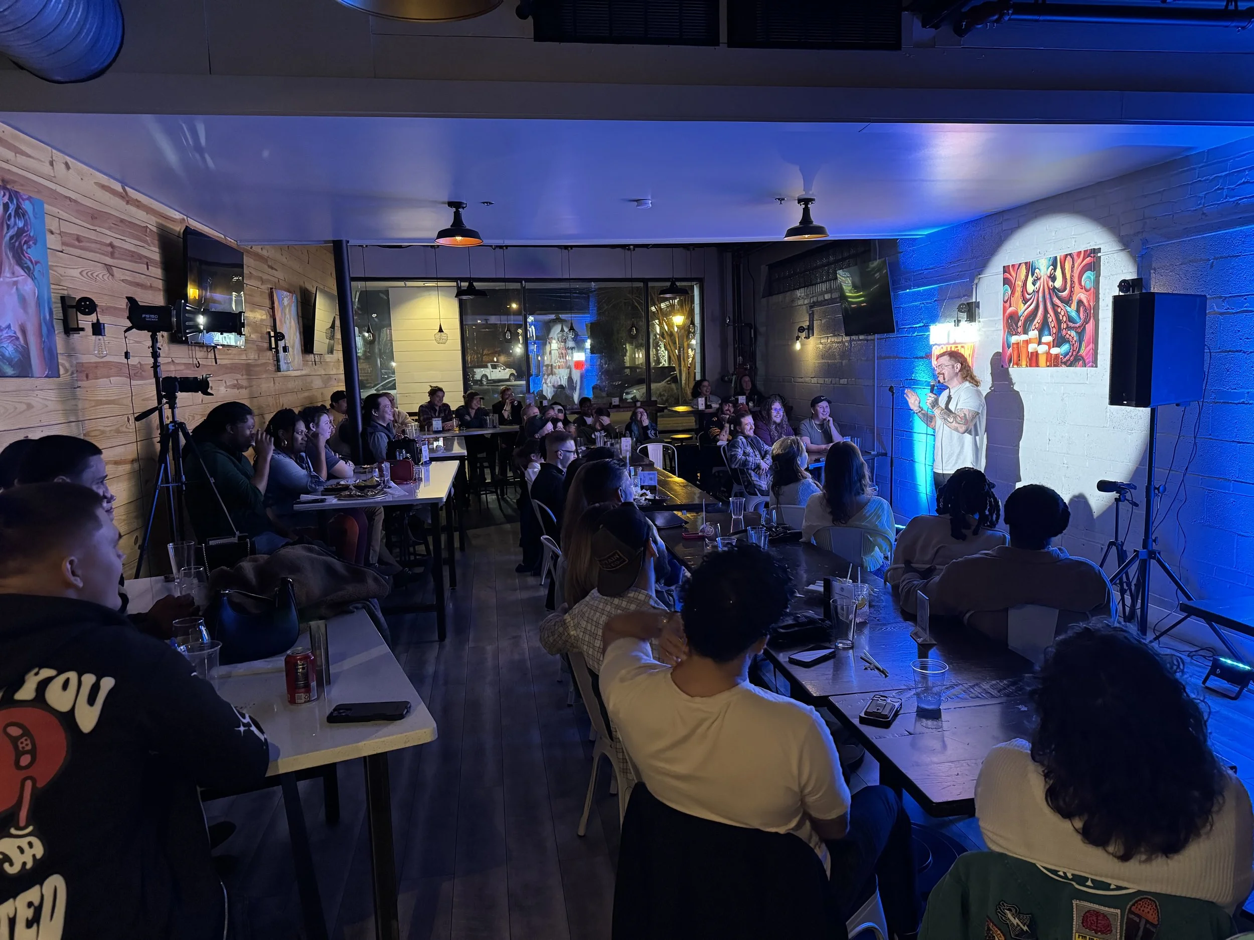 A comedy show is taking place in a cozy, dimly lit venue with an audience watching a performer on stage. The performer is a woman with glasses and tattoos, holding a microphone, illuminated by a spotlight. The audience is seated at tables, some with drinks, attentively watching the show. The venue has wooden and brick walls, and art and TVs decorated the space.