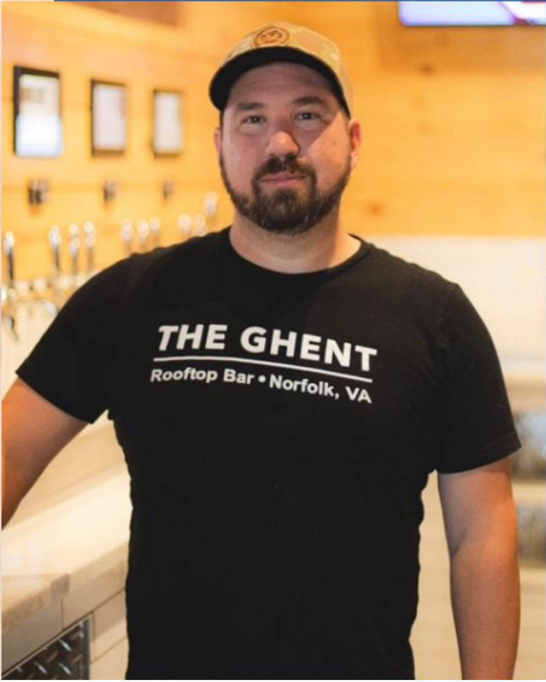 A man wearing a black T-shirt with white text reading "THE GHENT RoofTop Bar · Norfolk, VA" and a beige cap, standing in what appears to be a bar or restaurant with a wooden interior.