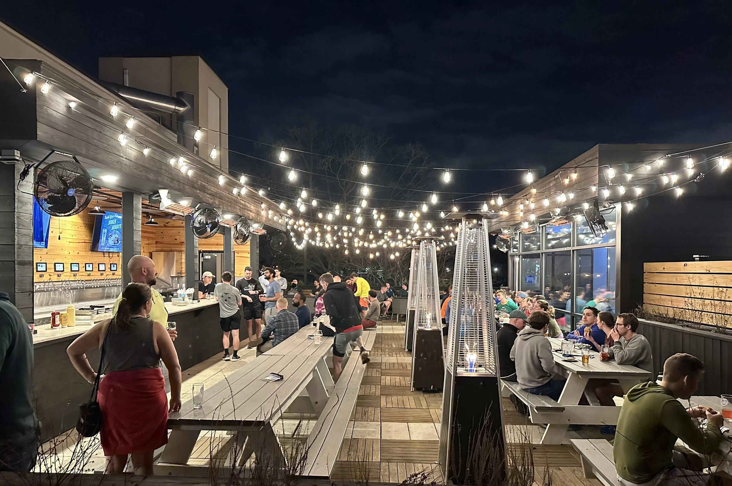 Outdoor rooftop bar at night with string lights, hanging out with people at tables and a bar counter, heaters, and fans.