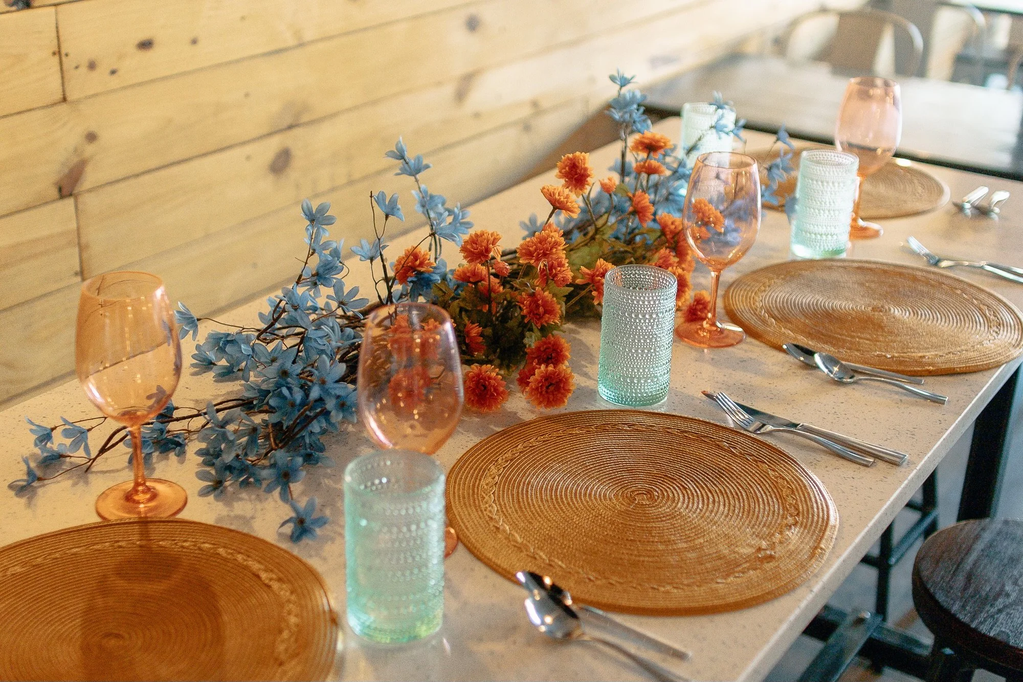 A dining table set with four orange textured placemats, four cocktail glasses with pinkish-orange tint, four textured light green drinking glasses, and a floral centerpiece of orange and blue flowers. The table has a light-colored surface and a wooden wall background.