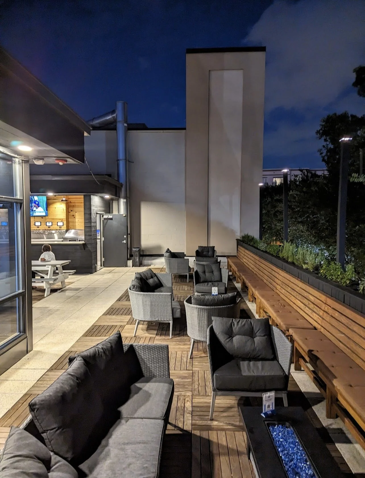 Outdoor patio with gray cushioned chairs, a long wooden bench, and a fire pit with blue glass stones. Some tables and a small child sitting at a picnic table are visible in the background. The patio is next to a building with a large chimney and a bar area with a TV.