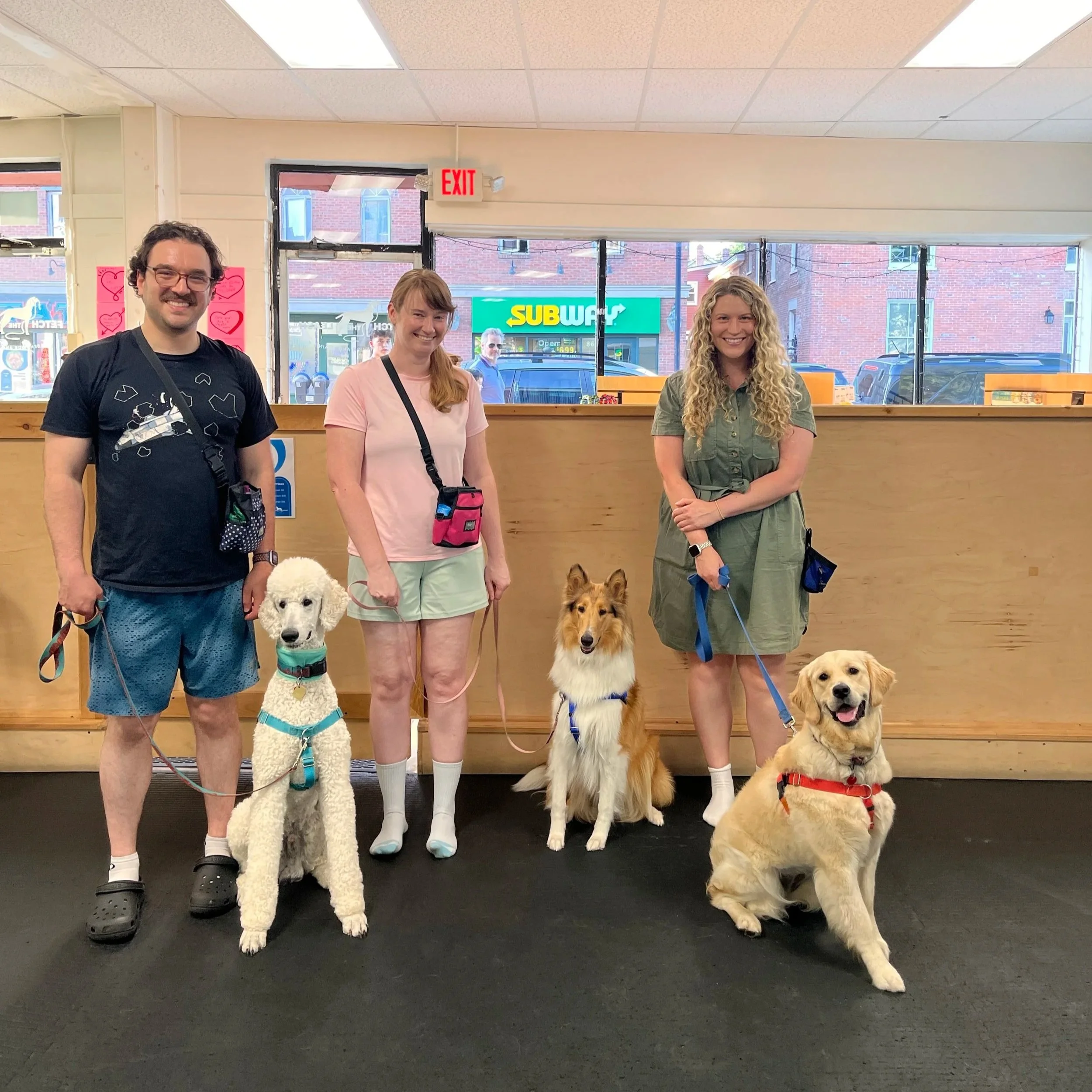 Three people and three dogs standing inside a store, with a Subway restaurant visible in the background through large windows.