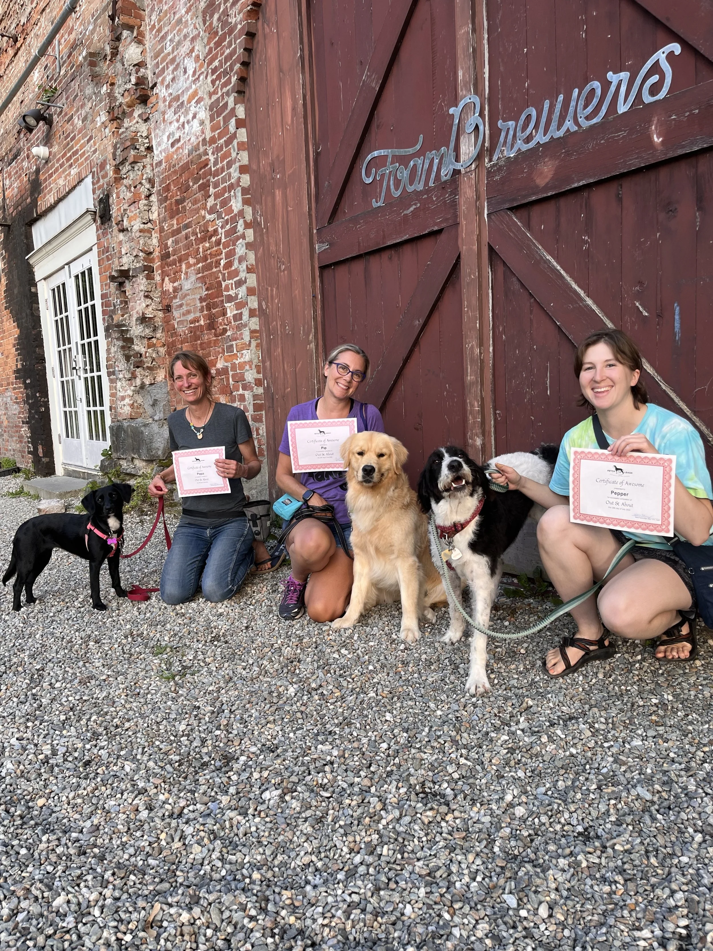 Three women with three dogs kneeling on gravel in front of a brick building with a large red wooden door, holding certificates of achievement and smiling.