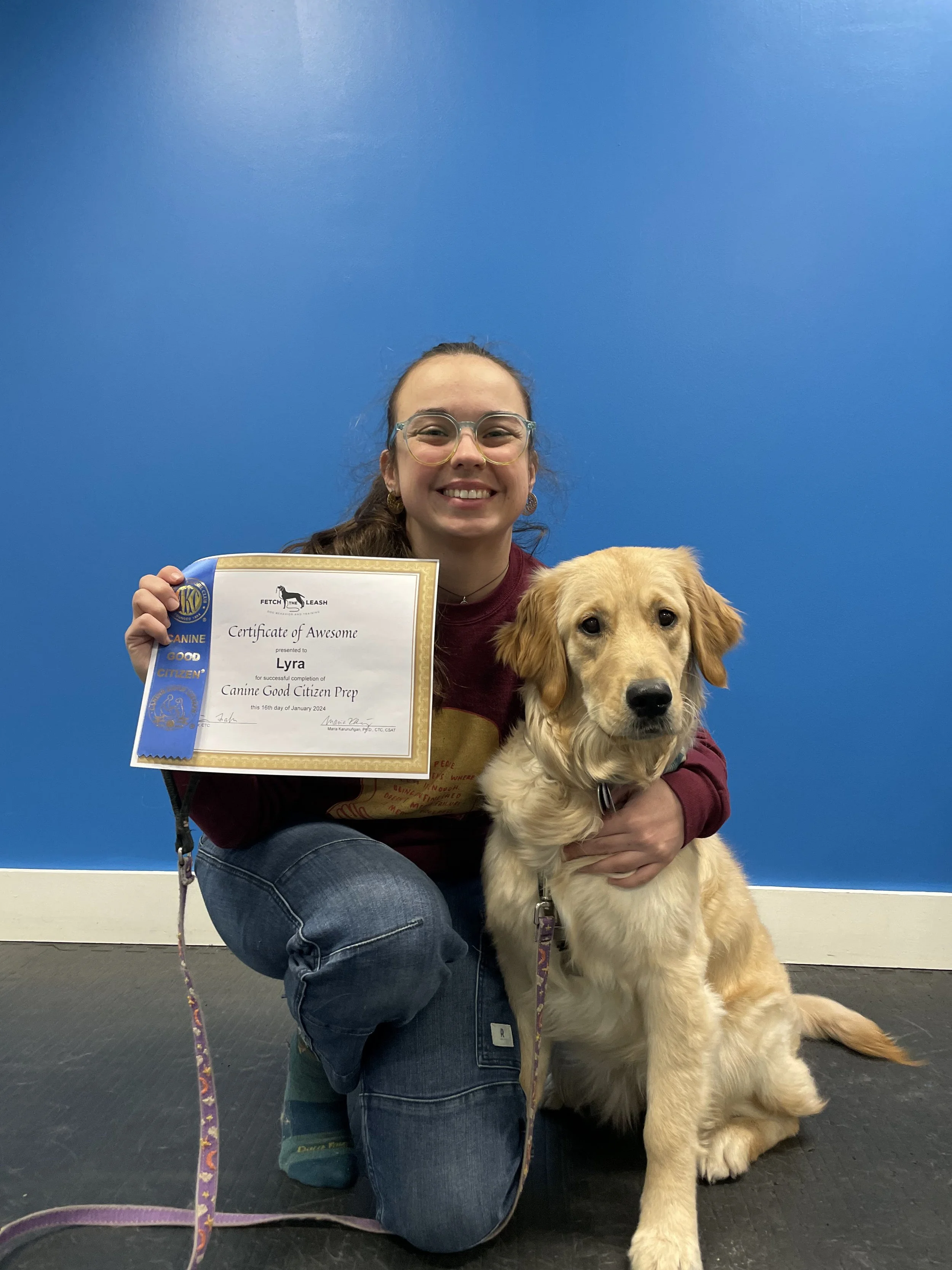 Young woman with glasses kneeling beside a golden retriever, holding a certificate of achievement, against a blue wall.