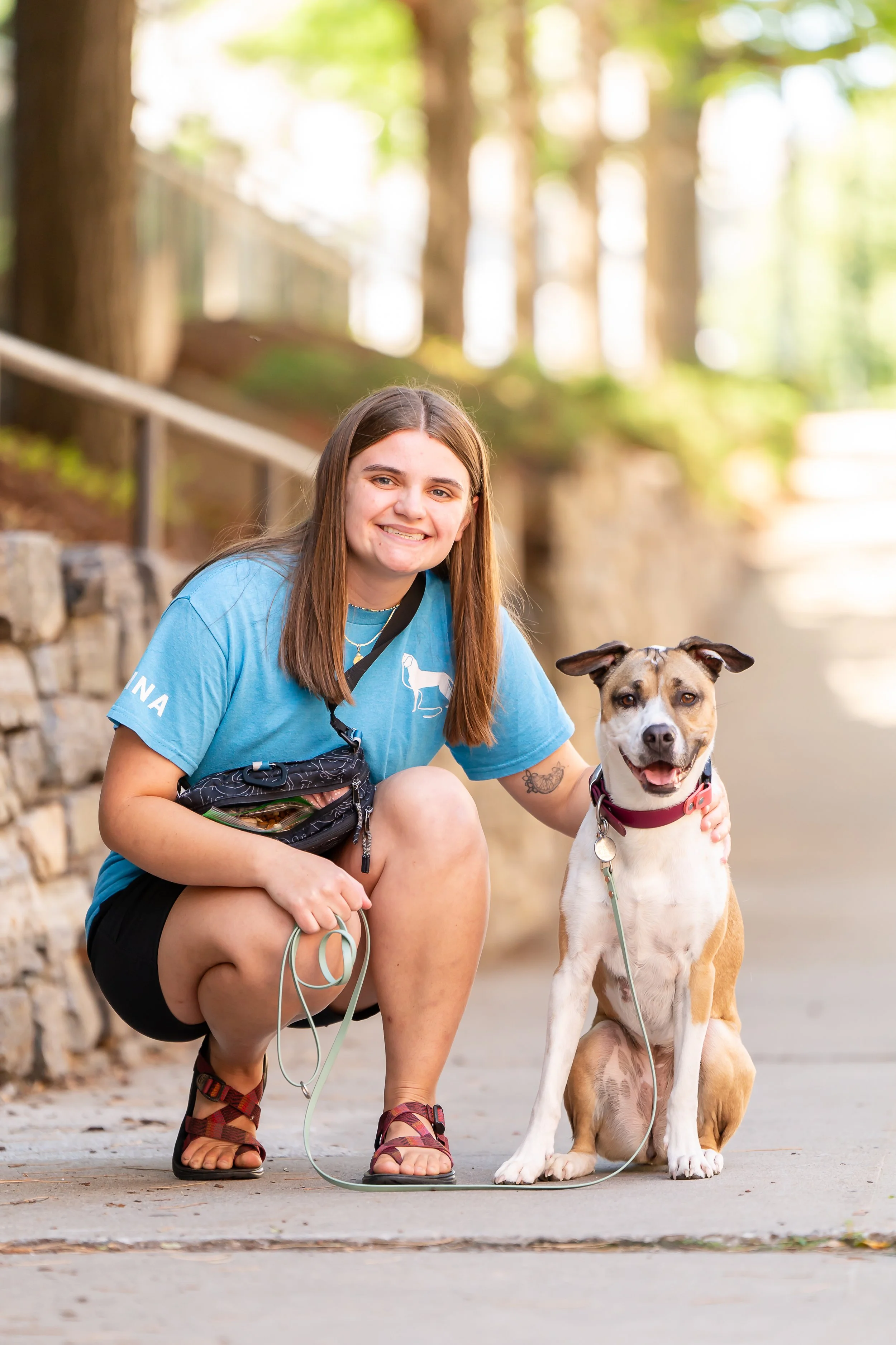 A young woman with shoulder-length brown hair crouching next to a smiling dog on a leash outdoors in a park with trees in the background.