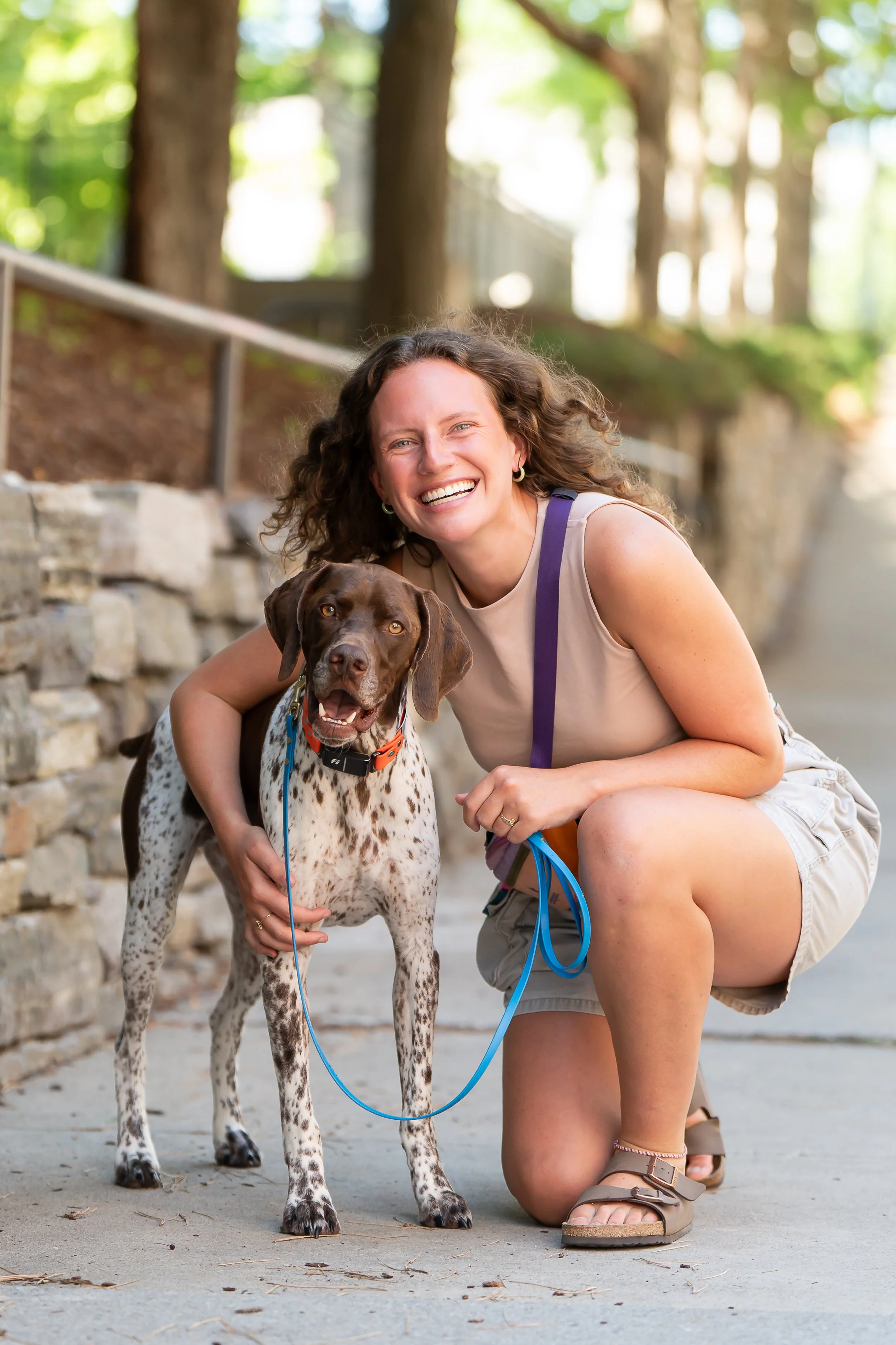A woman with curly brown hair, wearing a beige sleeveless top and khaki shorts, kneeling on a sidewalk with a brown and white spotted dog, both smiling happy, in a park with trees and a stone wall in the background.