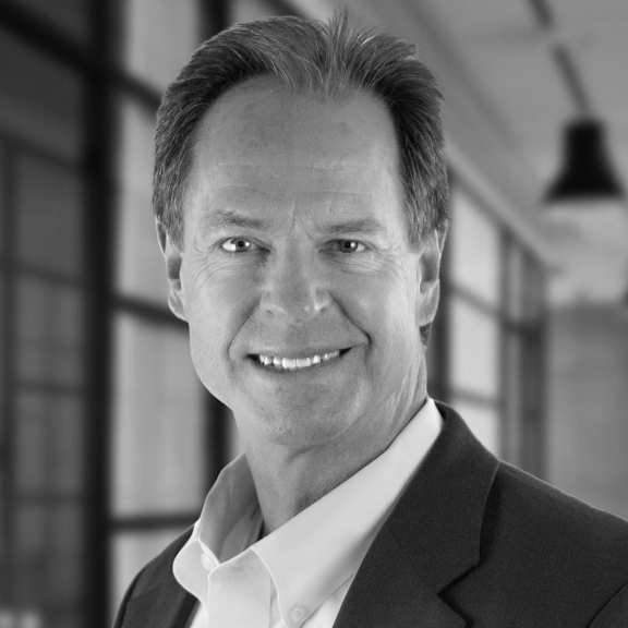 Portrait of Realtor Bobby Mclain in a suit smiling in an indoor setting with large windows in the background.