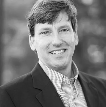 Realtor Rex Carter, with light-colored hair wearing a dark blazer and checkered shirt, smiling outdoors.