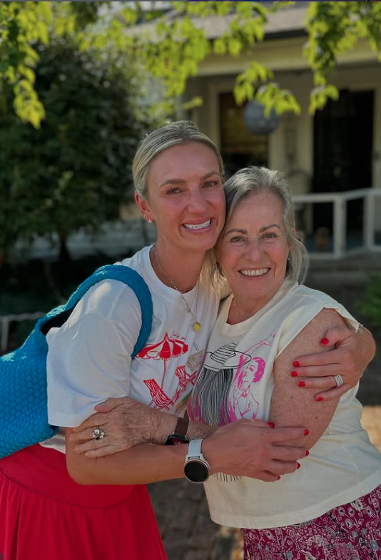 Two women smiling and hugging outdoors in front of trees and a house.