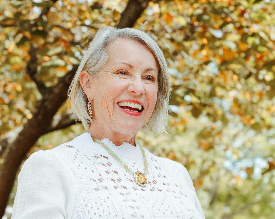 Realtor Lil Glenn with short gray hair smiling outdoors, wearing a white lace blouse, a necklace, and earrings, with a tree with autumn leaves in the background.