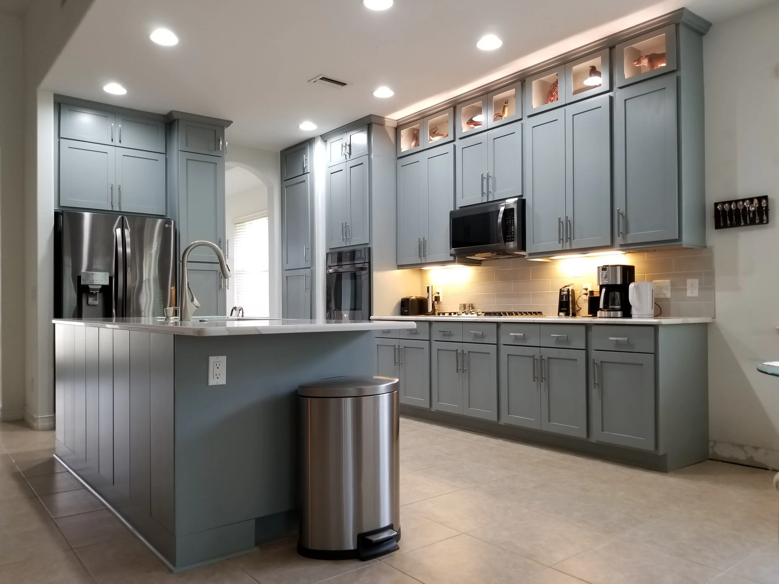Modern kitchen with gray cabinets, stainless steel refrigerator, black microwave, coffee maker, and white countertop with island.