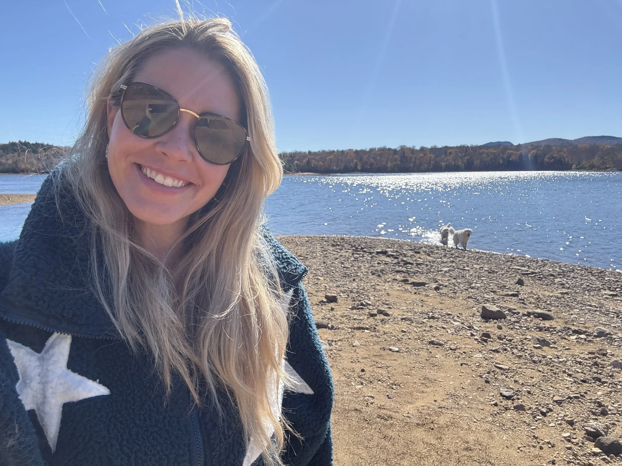 Smiling woman wearing sunglasses taking a selfie near a lake with two white dogs, sunny day with clear blue sky and distant hills.