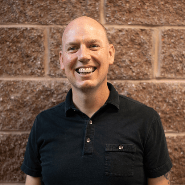 Smiling man with a shaved head in a black shirt standing against a brick wall background.