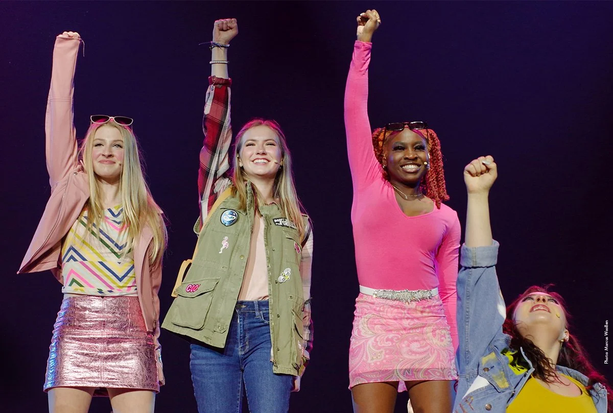 Stage adaptation of Mean Girls, with four girls raising their arms in the air.