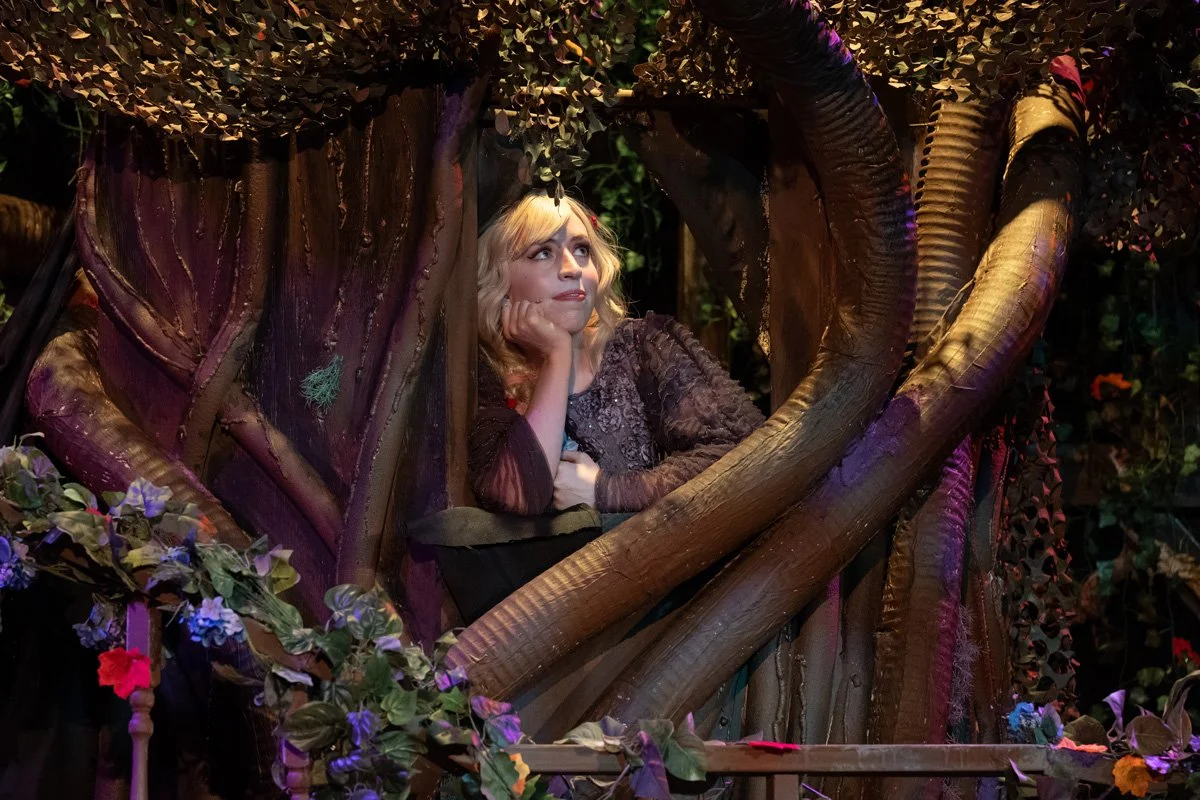 Woman with blonde hair sitting in a large tree in theater performance.