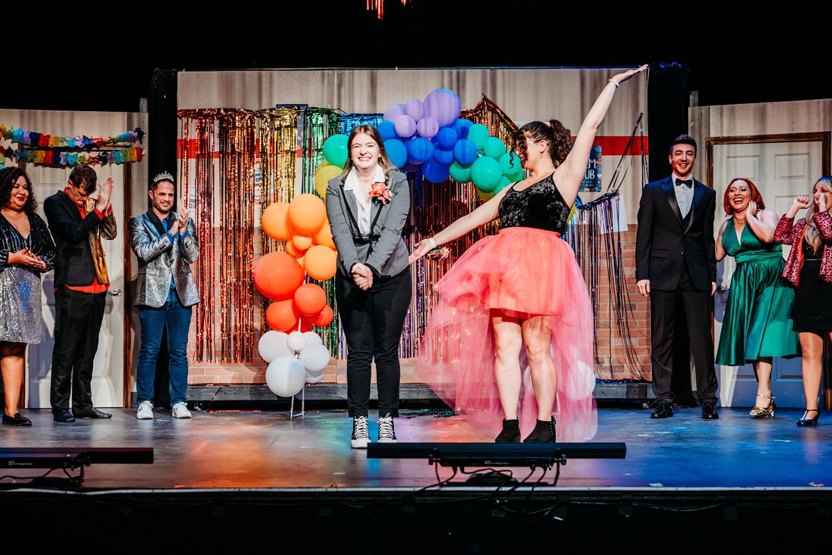 Group of people on a stage at a celebration or event, with some dressed in formal attire and one woman wearing a pink tulle skirt and black top, holding her arm up. Colorful balloons and decorations are in the background.