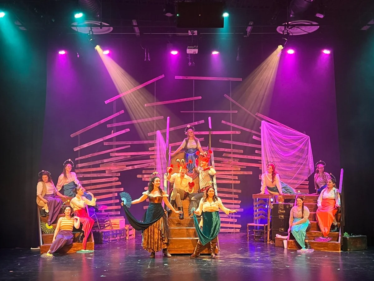 Stage scene from a theatrical play with multiple actors dressed in period costumes, colorful lighting, and a wooden set design.