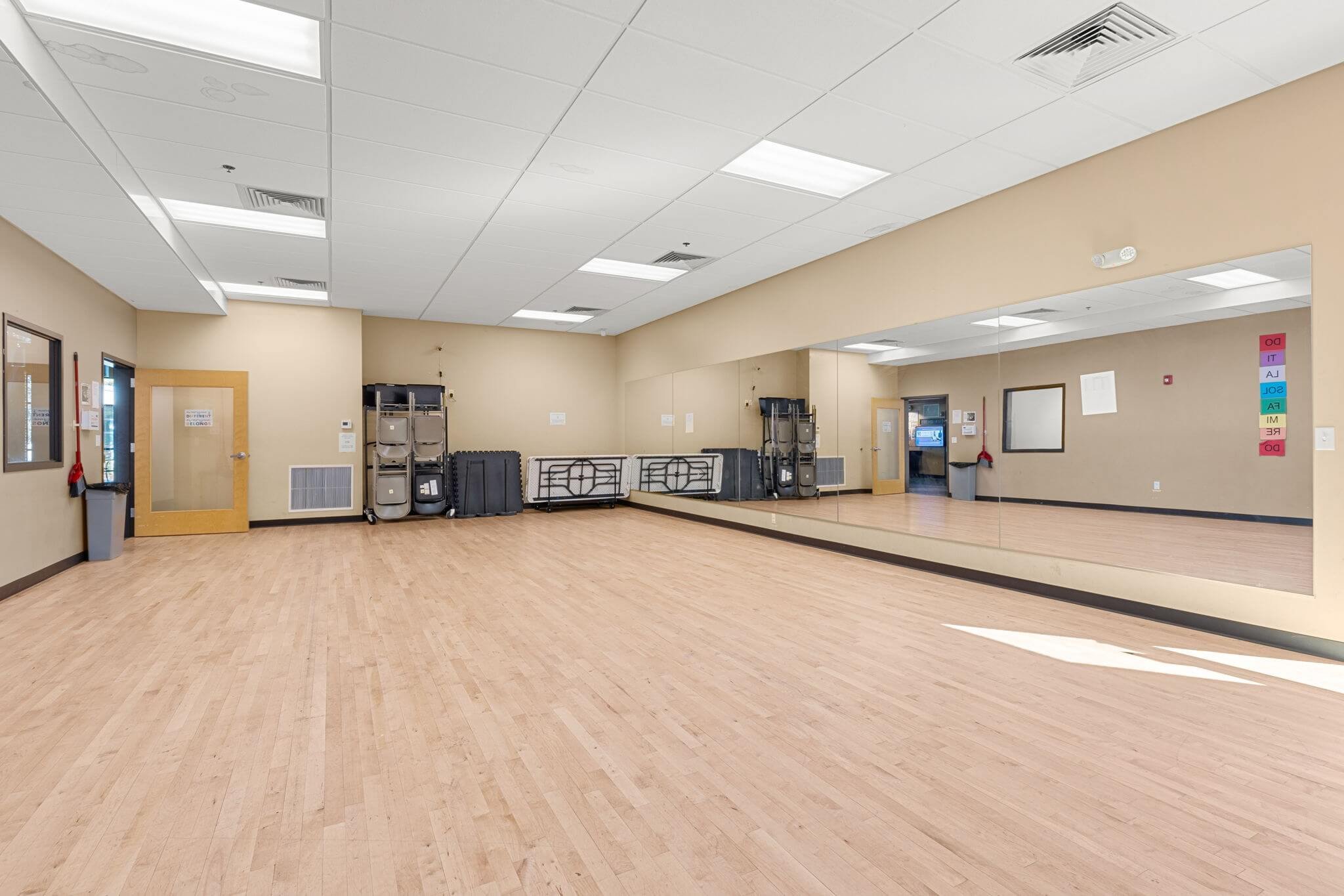 Empty dance studio with large mirror, wood flooring, folding chairs, and a door with a window.