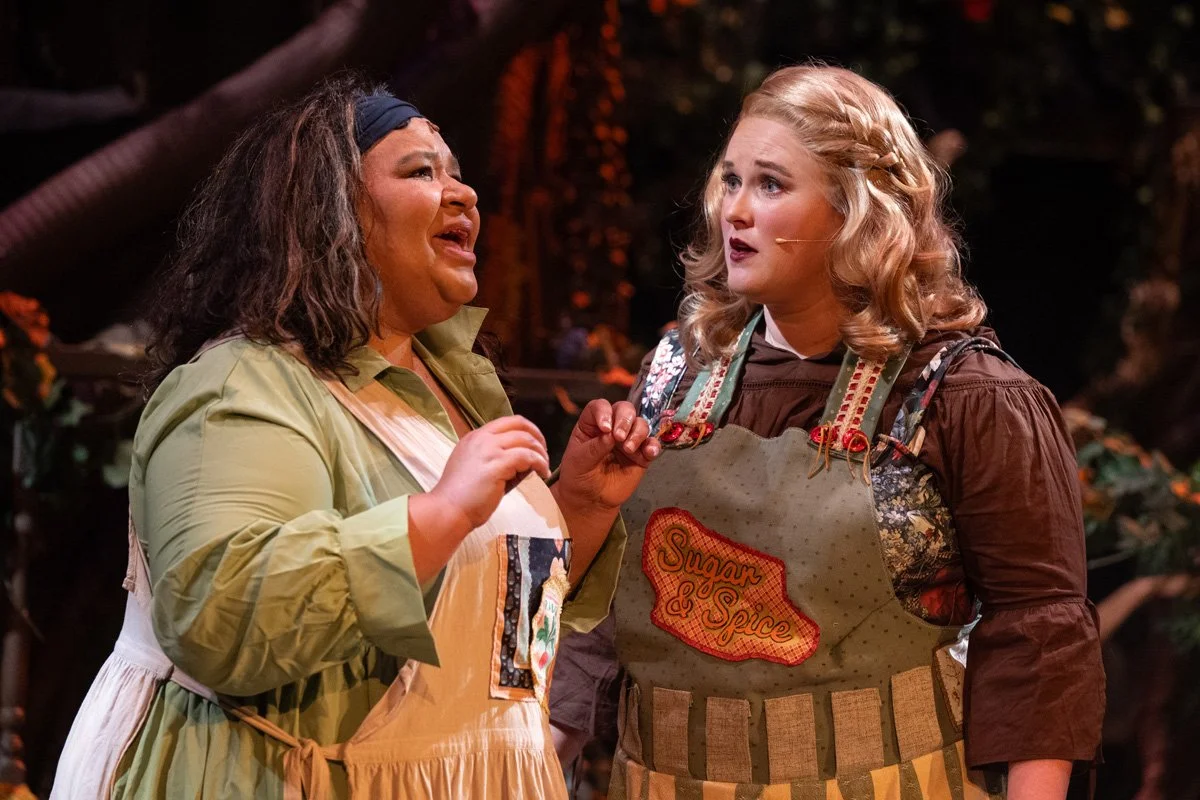 Two women, one with dark hair and the other with blonde hair, wearing aprons and talking to each other on a stage with a dark background. 