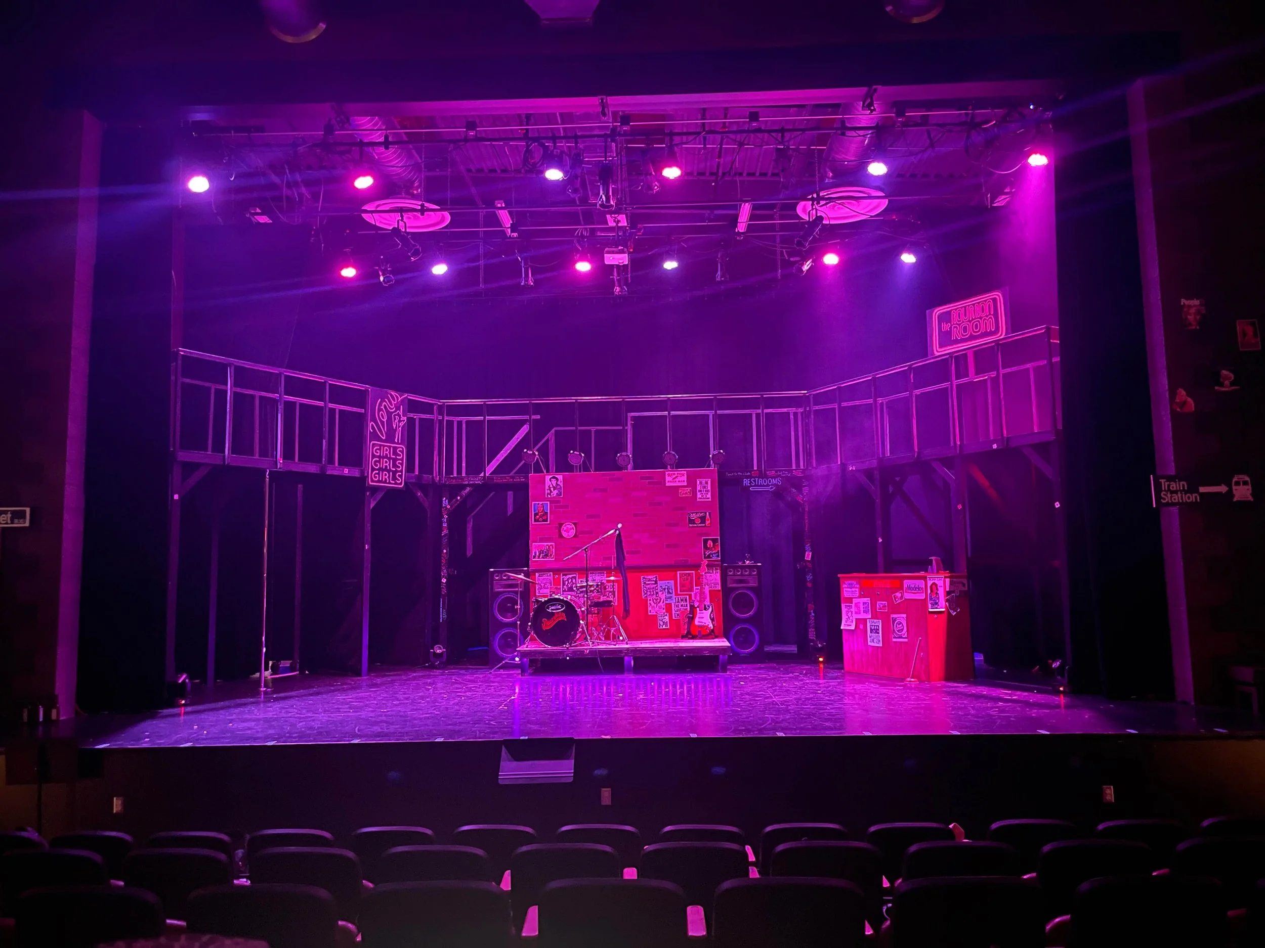 Stage setup for a performance with purple and pink lighting, musical instruments including drums, microphones, and speakers, decorated with posters and signs, with an elevated balcony and seating in front.