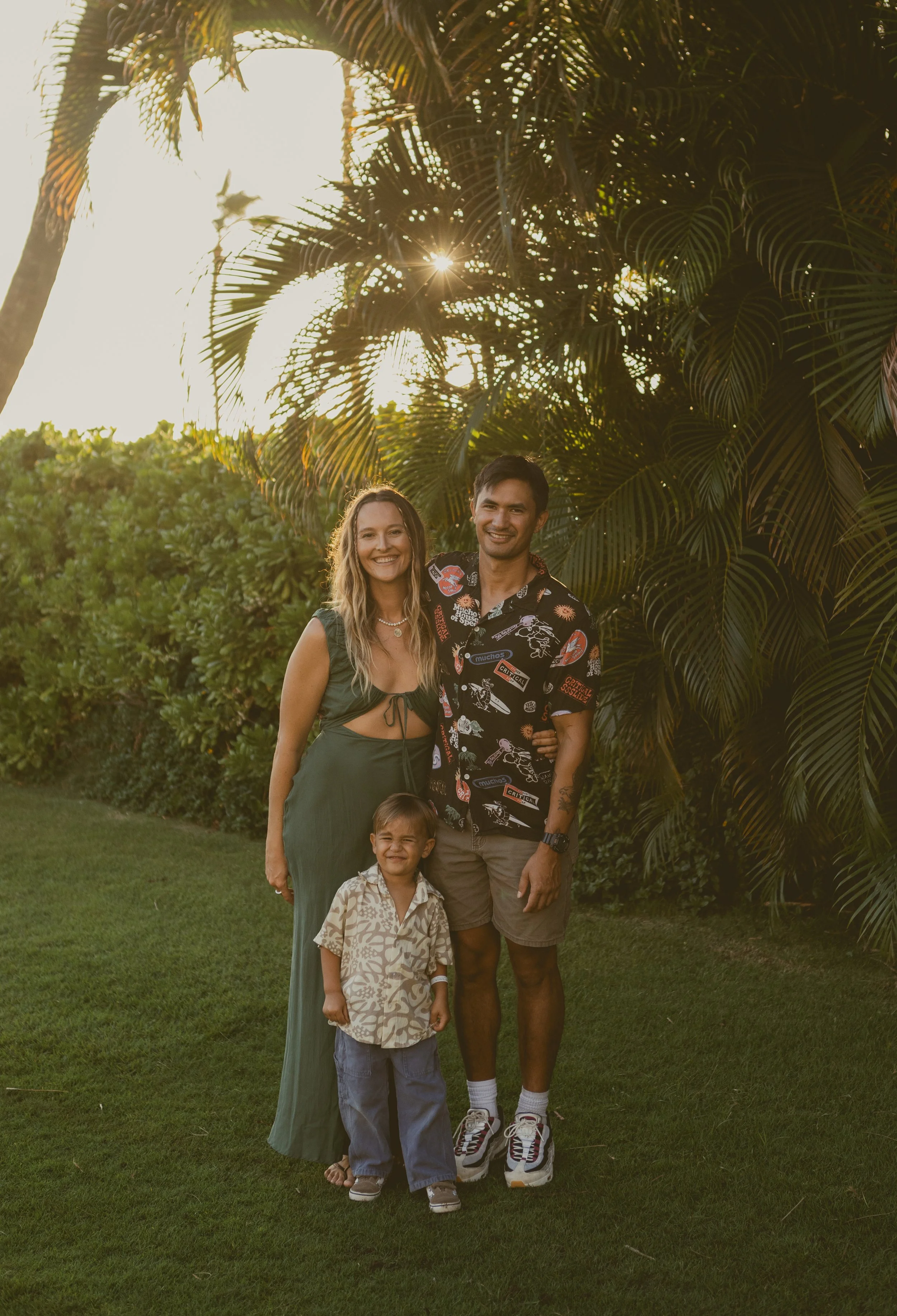 Maui Super 8mm videographer. family of three standing outdoors on green grass, smiling, with lush tropical plants and the sun shining behind large palm leaves.