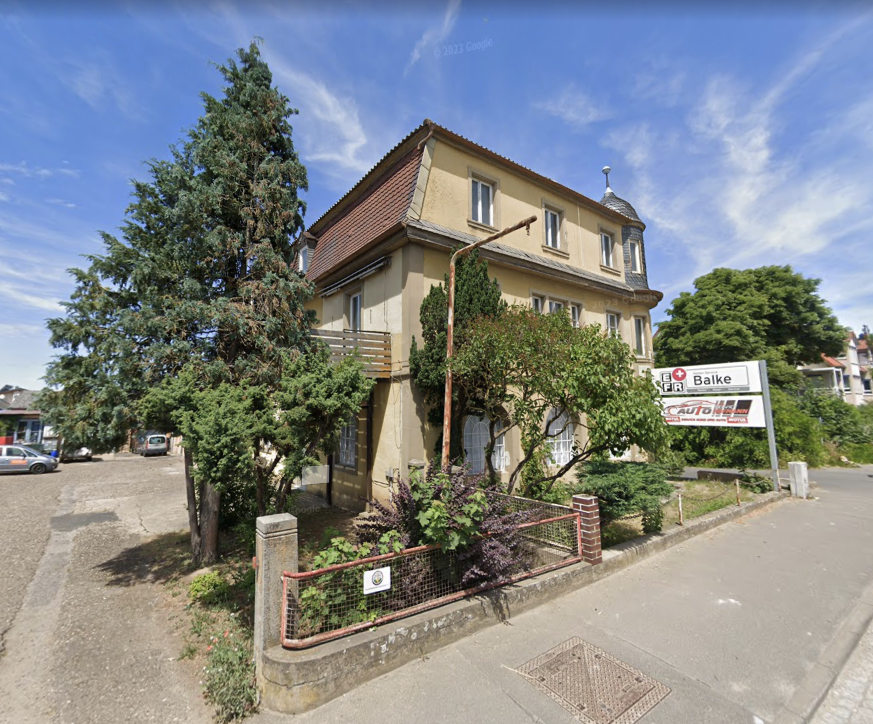 Ein mehrstöckiges, altes Wohnhaus mit gelber Fassade und roten Ziegel-Dach, umgeben von Bäumen und Sträuchern, mit einem Parkplatz im Vordergrund und einem Schild mit der Aufschrift 'Balke'.
