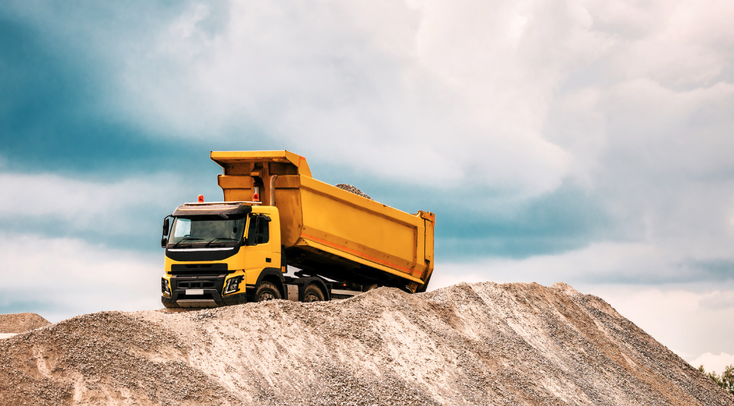 A yellow dump truck on a mound of gravel or dirt outdoors with a cloudy sky in the background.