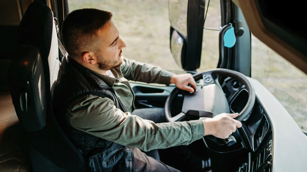 A man sitting in the driver's seat of a large truck, holding the steering wheel with both hands, looking out the window.