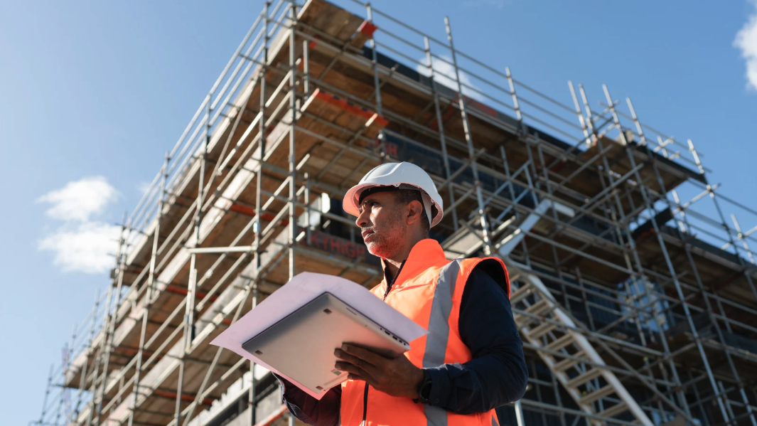 Construction worker wearing a white hard hat and orange safety vest holding a tablet, standing in front of a scaffolding-covered building under a blue sky.