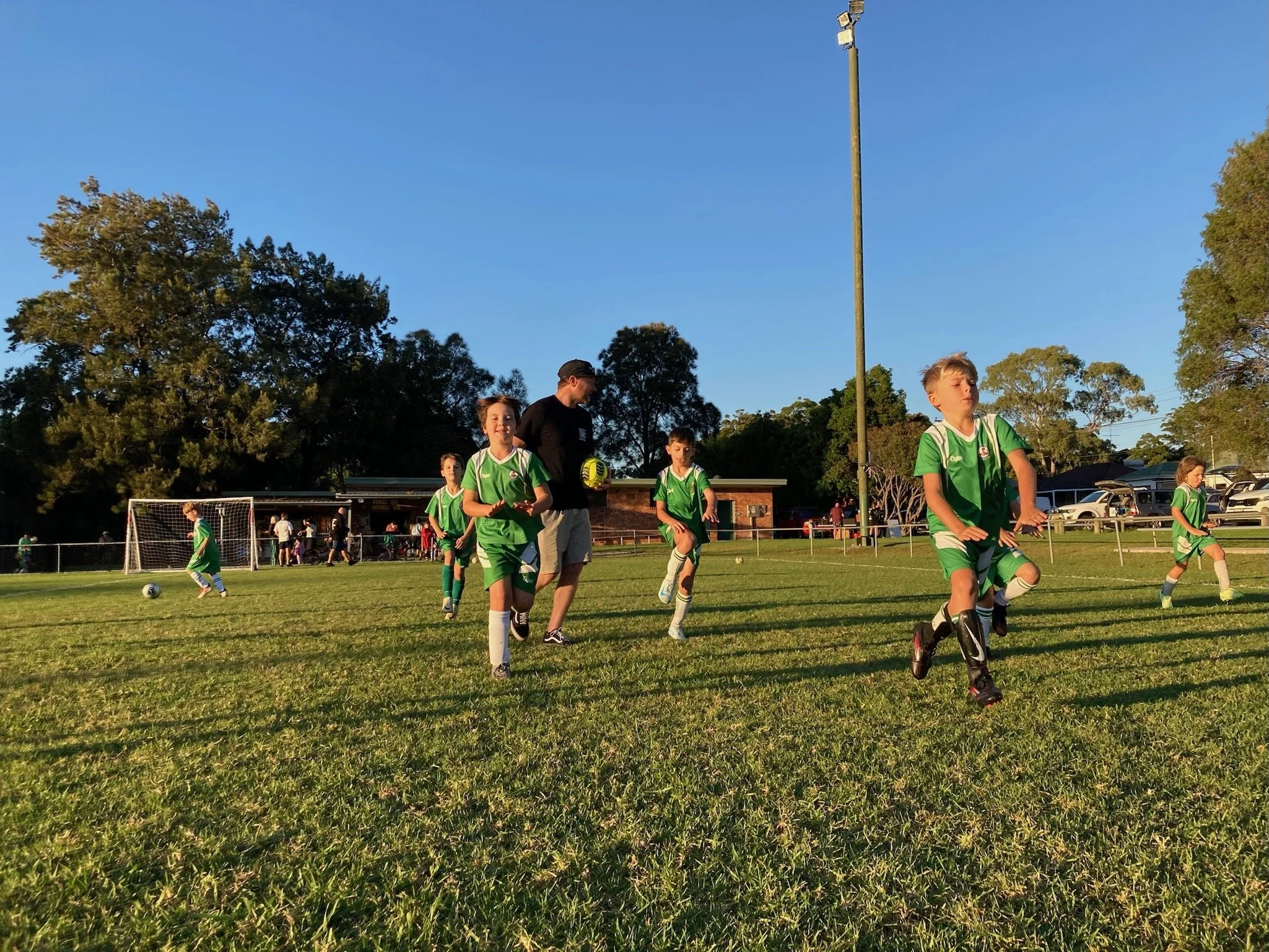 Children in green soccer uniforms jogging on a soccer field while a coach in black shirt and shorts holds a soccer ball, with goalposts and trees in the background during sunset.
