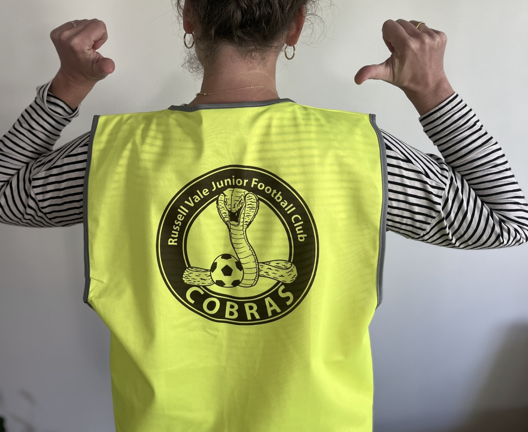 A woman wearing a yellow safety vest with the logo for Russell Vale Junior Football Club COBRAS and flexing her arms. She has earrings, a necklace, and a striped long-sleeve shirt underneath the vest. The background is plain and light-colored.