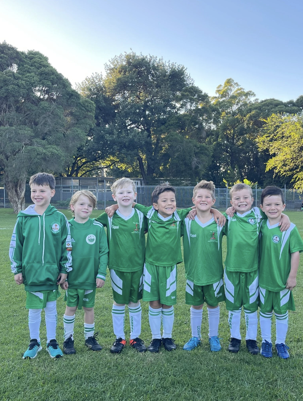 Seven young boys in green soccer uniforms standing together with arms around each other on a grassy field, smiling. Trees and a fence are in the background under a clear blue sky.