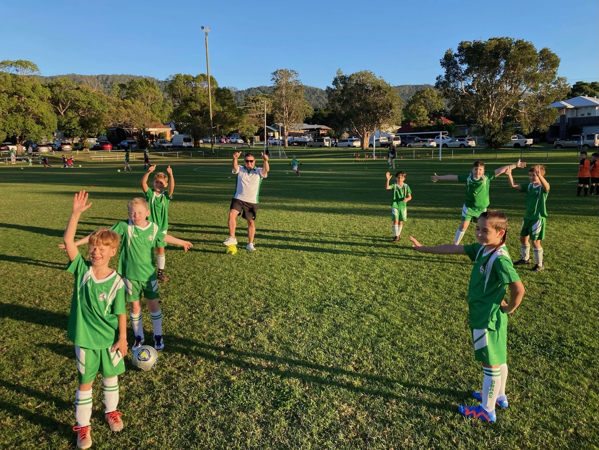 Kids in green soccer uniforms standing on a grassy field with a coach, smiling and waving, with other children and trees in the background under a clear blue sky.