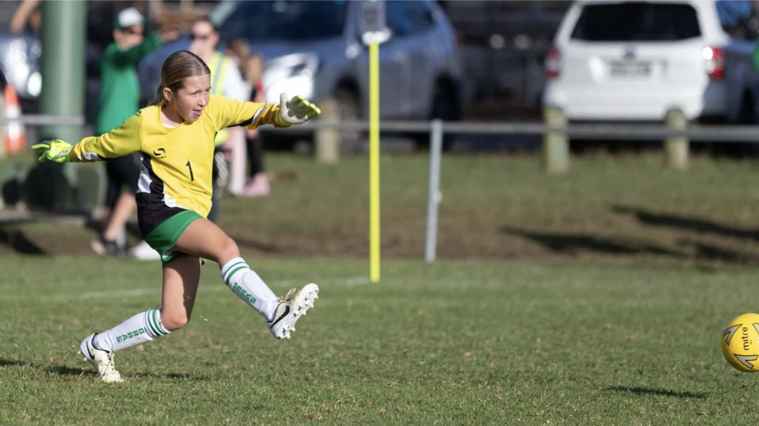 A young girl in a yellow goalkeeper jersey, green shorts, and white socks with green stripes, is playing soccer outdoors, kicking a yellow soccer ball on a grassy field during a game.