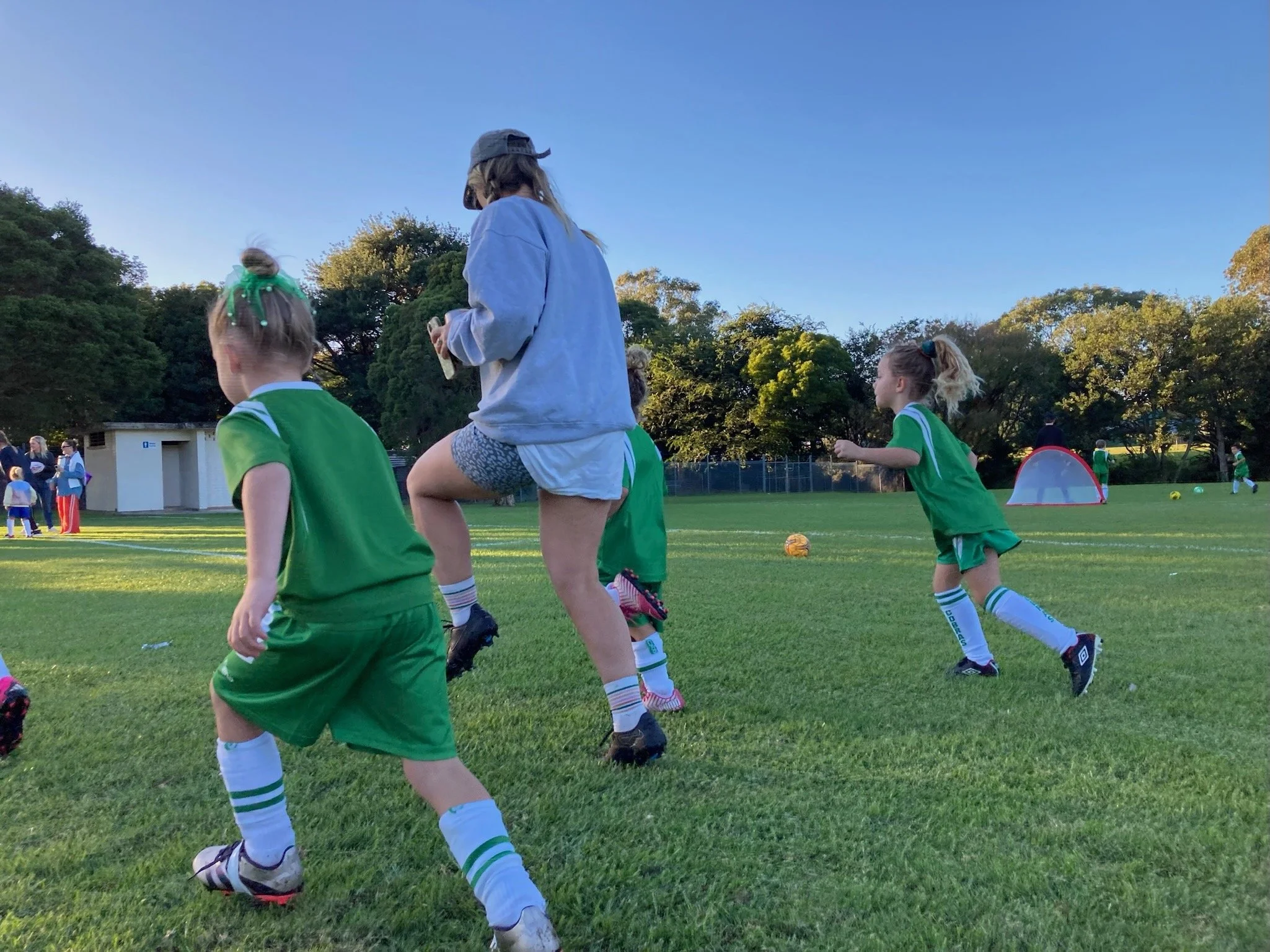 Young girls playing soccer on a green field during the daytime, with children and adults in the background, and trees and a small building nearby.